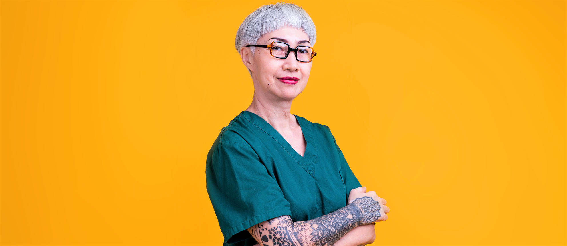 Eine ältere Frau mit Tattoos und kurzen Haaren posiert lächelnd in einer typischen Berufsbekleidung für Pfleger*innen vor einem einfarbigen gelben Hintergrund.