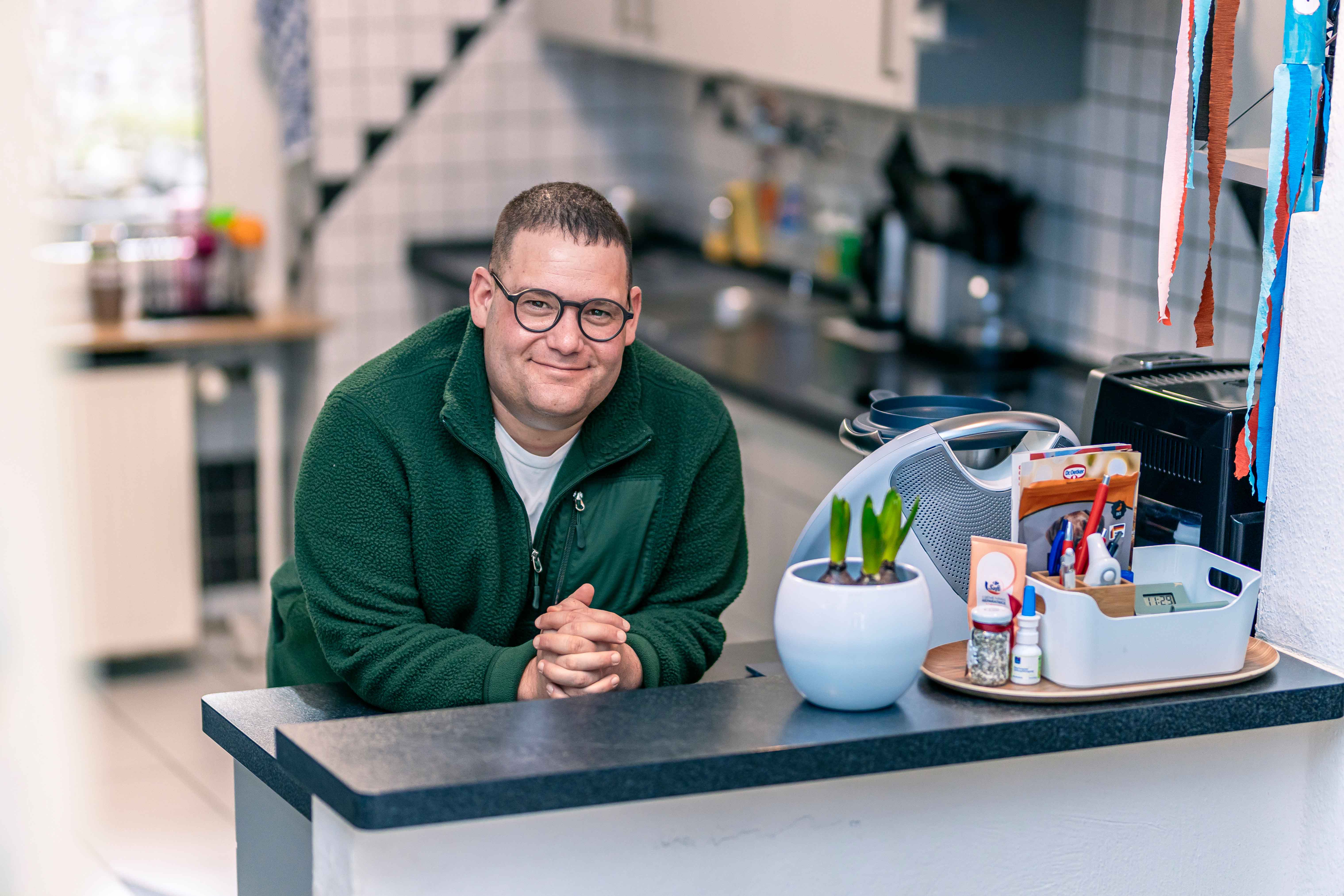 Ein Mann mit grünem Pulli und Brille lehnt in einer Küche an einem Tresen und lächelt herzlich in die Kamera.