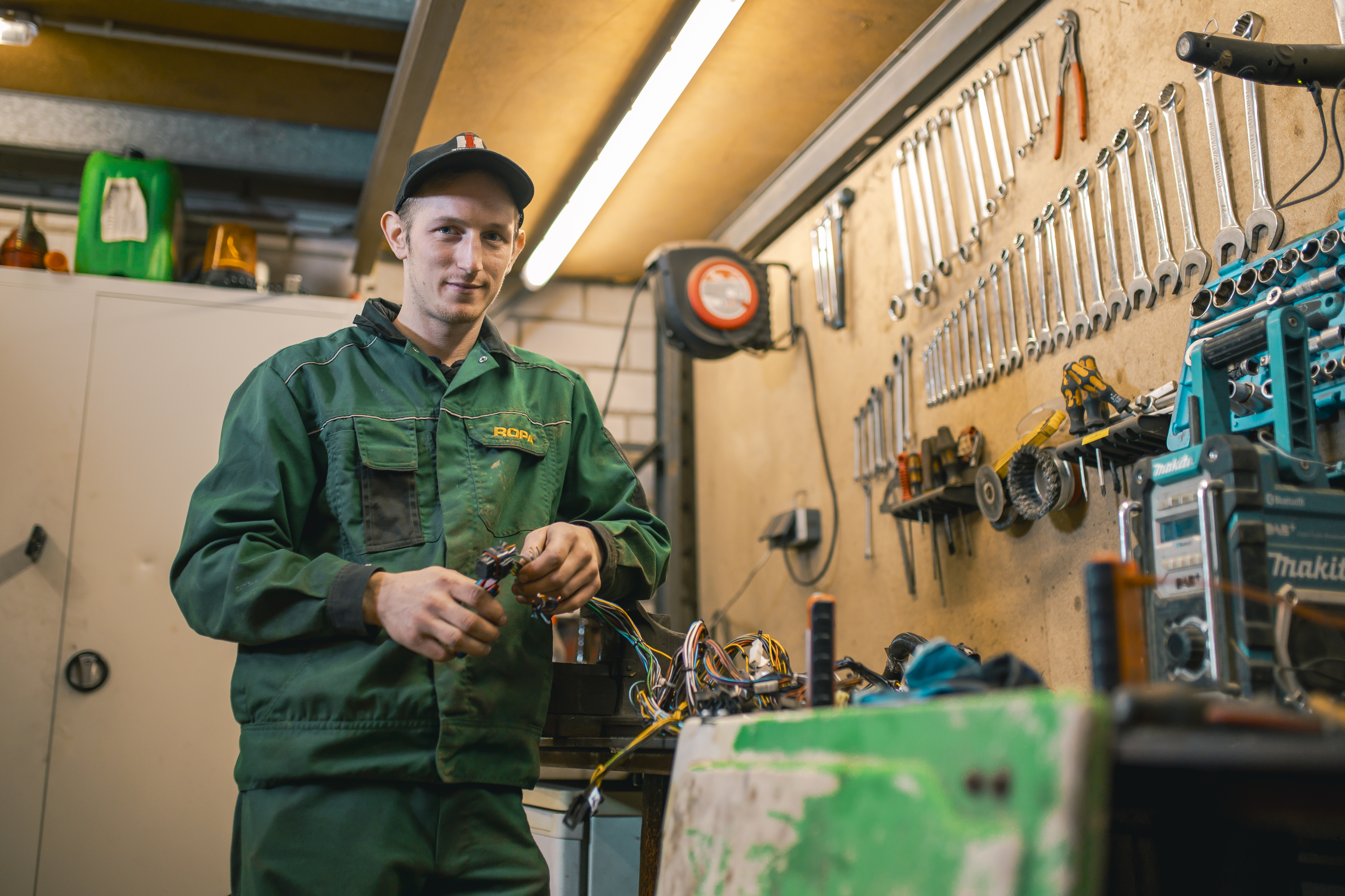 Ein junger Mann in Arbeitskleidung und mit Kappe  in einer Werkstatt. An der Wand hängen Schraubenschlüssel, er hat Kabel in der Hand und  blickt leicht lächelnd in die Kamera.