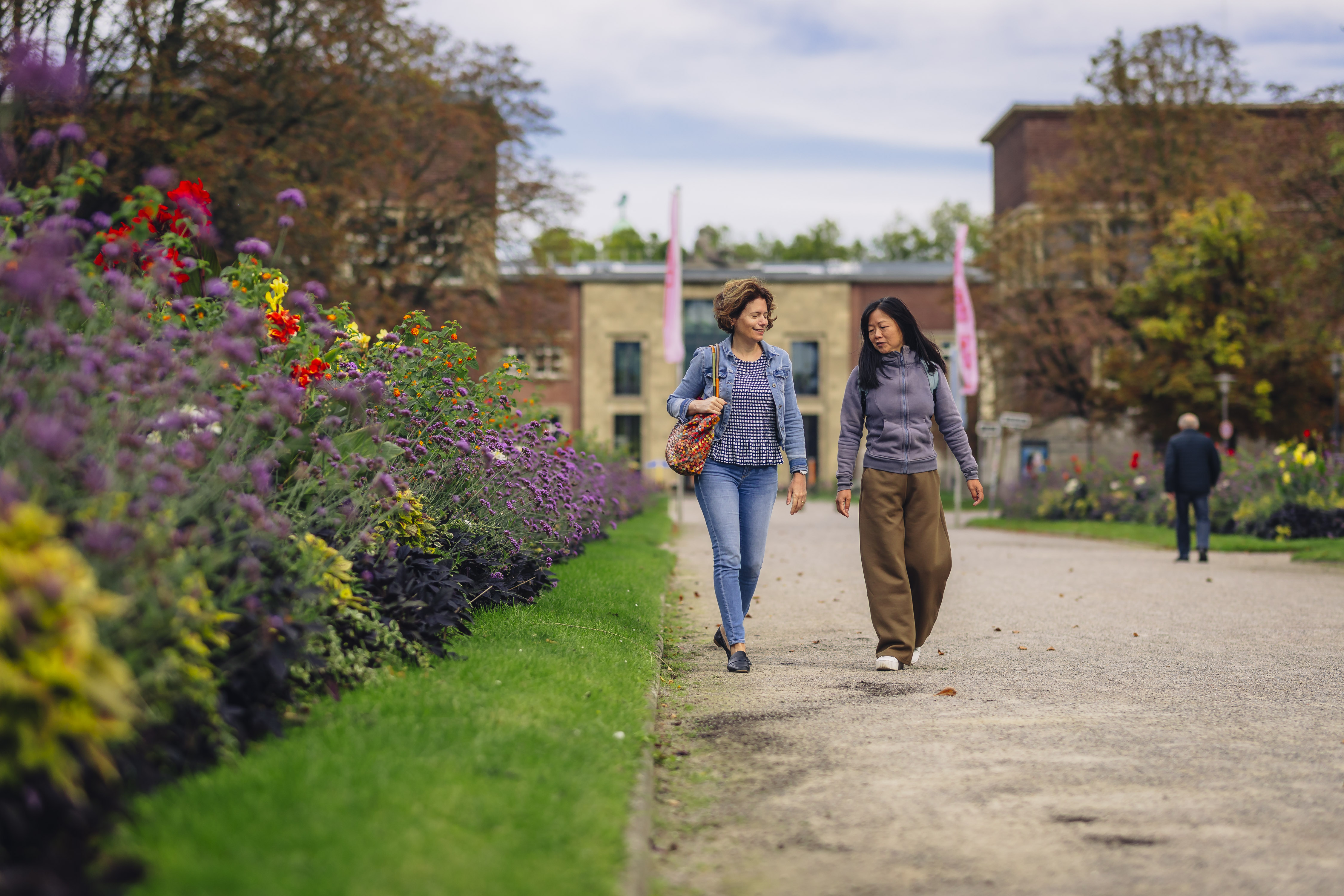 Zwei Frauen laufen auf einem sandigen Weg durch den Düsseldorfer Ehrenhof, vorbei an bunten Blumenbeeten. Im Hintergrund ist der Kunstpalast zu sehen.