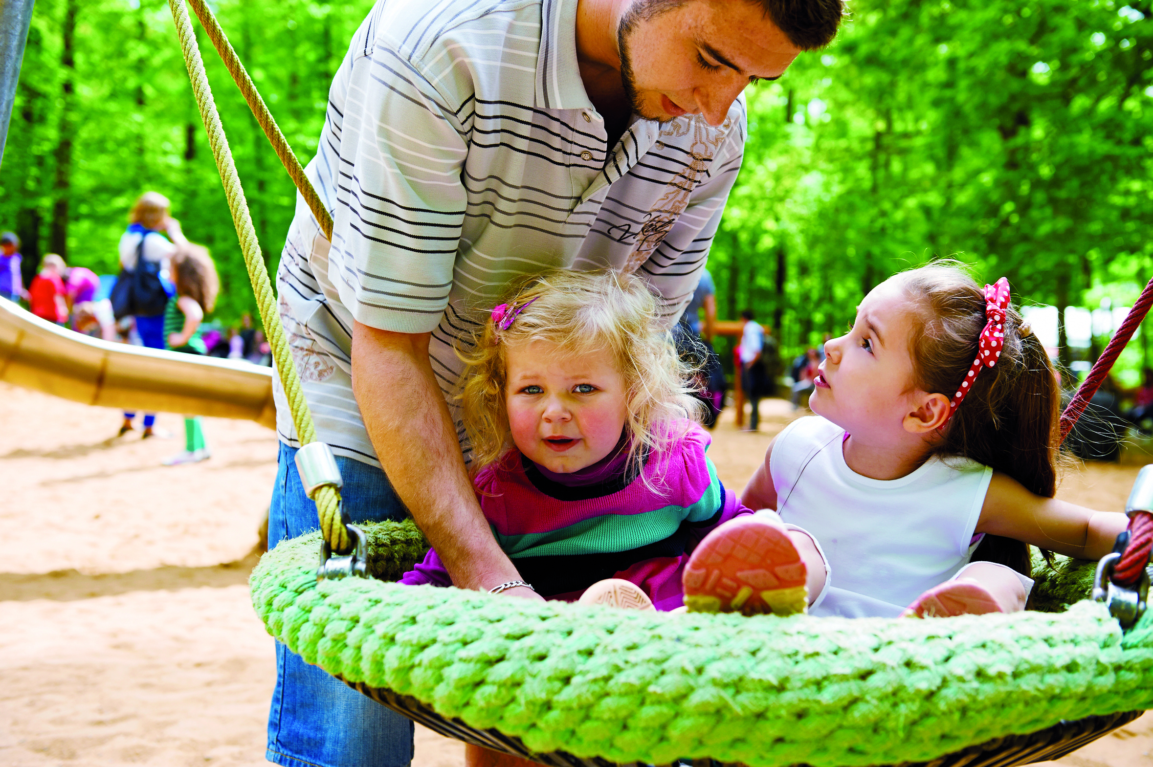 Ein junger Erzieher setzt zwei kleine Mädchen in einer Kita auf eine Nest-Schaukel.