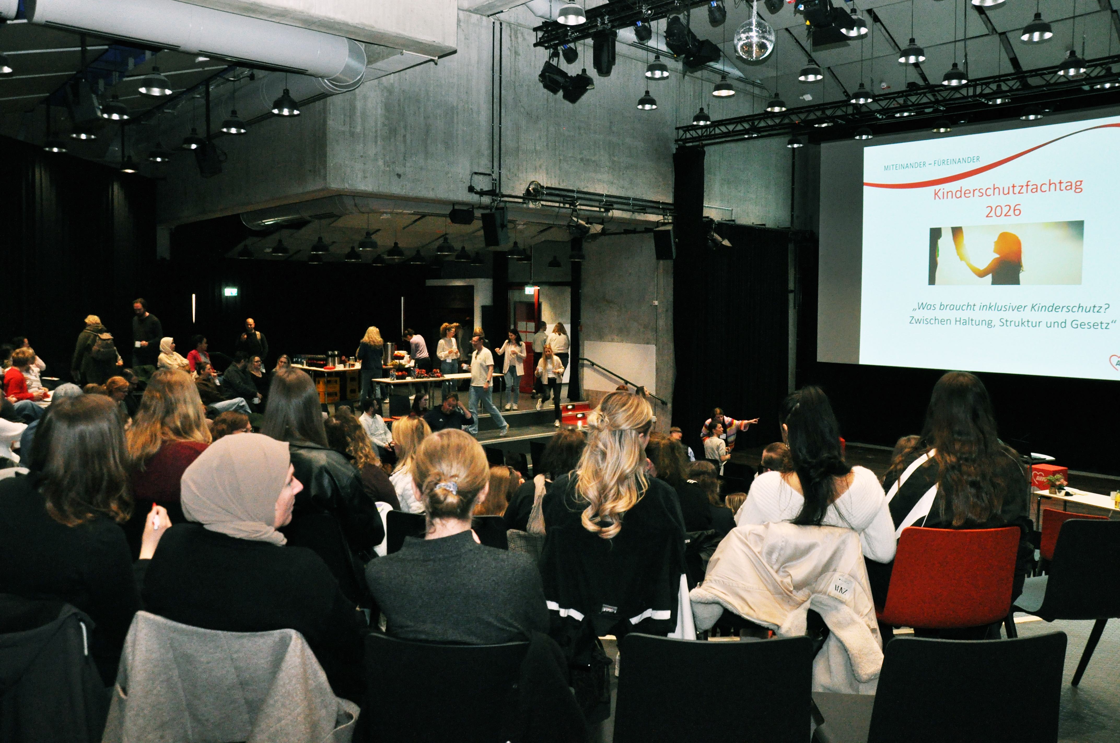 Blick von hinten auf eine Bühne, über viele Hinterköpfe von Zuschauenden hinweg. An die Wand ist eine Folie gebeamt auf  der steht: Kinderschutzfachtag 2026