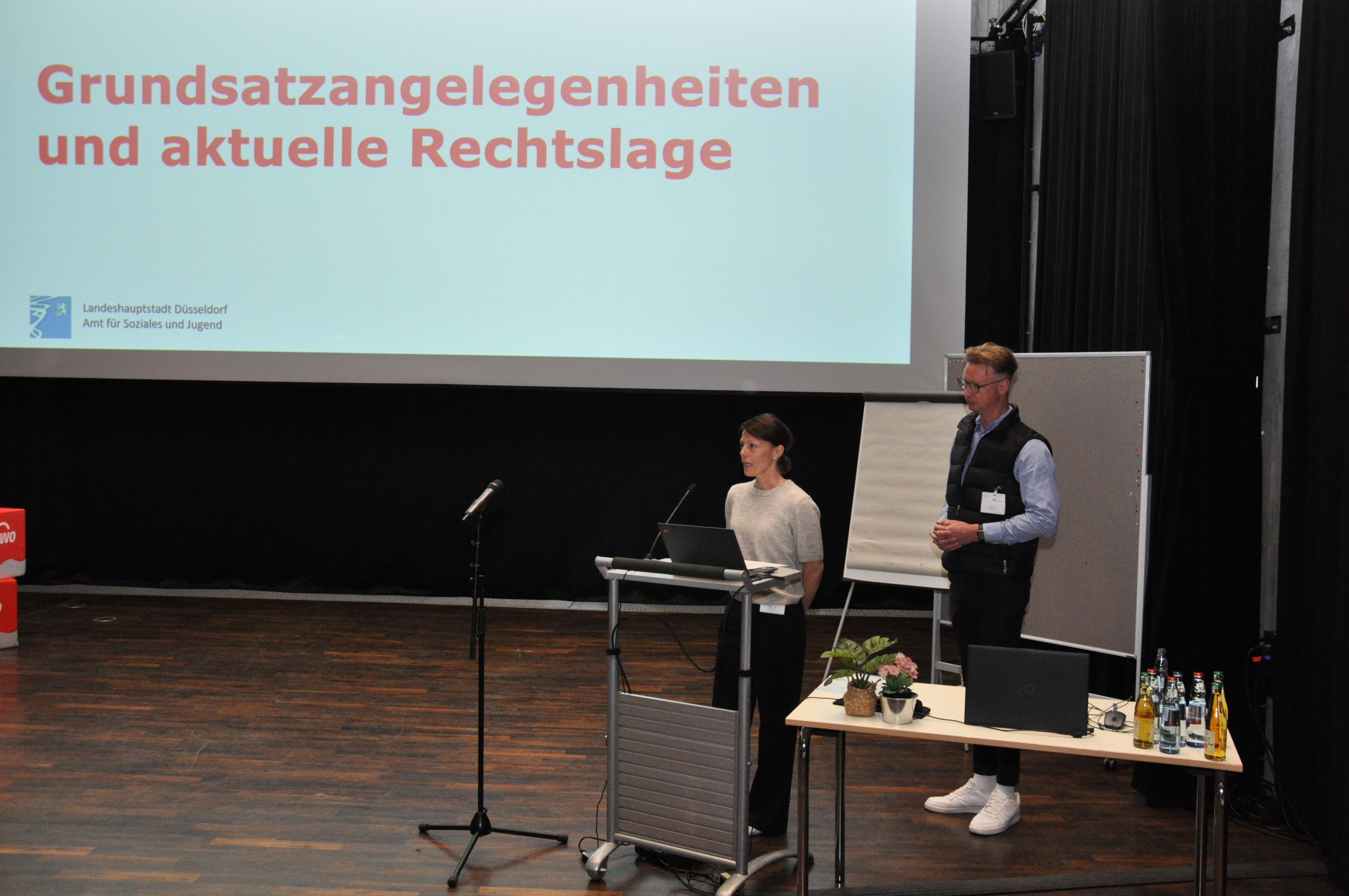Blick auf die Bühne, die beiden Referent*innen sprechen, hinter ihnen ist eine Folie projiziert, auf der steht: Grundsatzangelegenheiten und aktuelle Rechtslage
