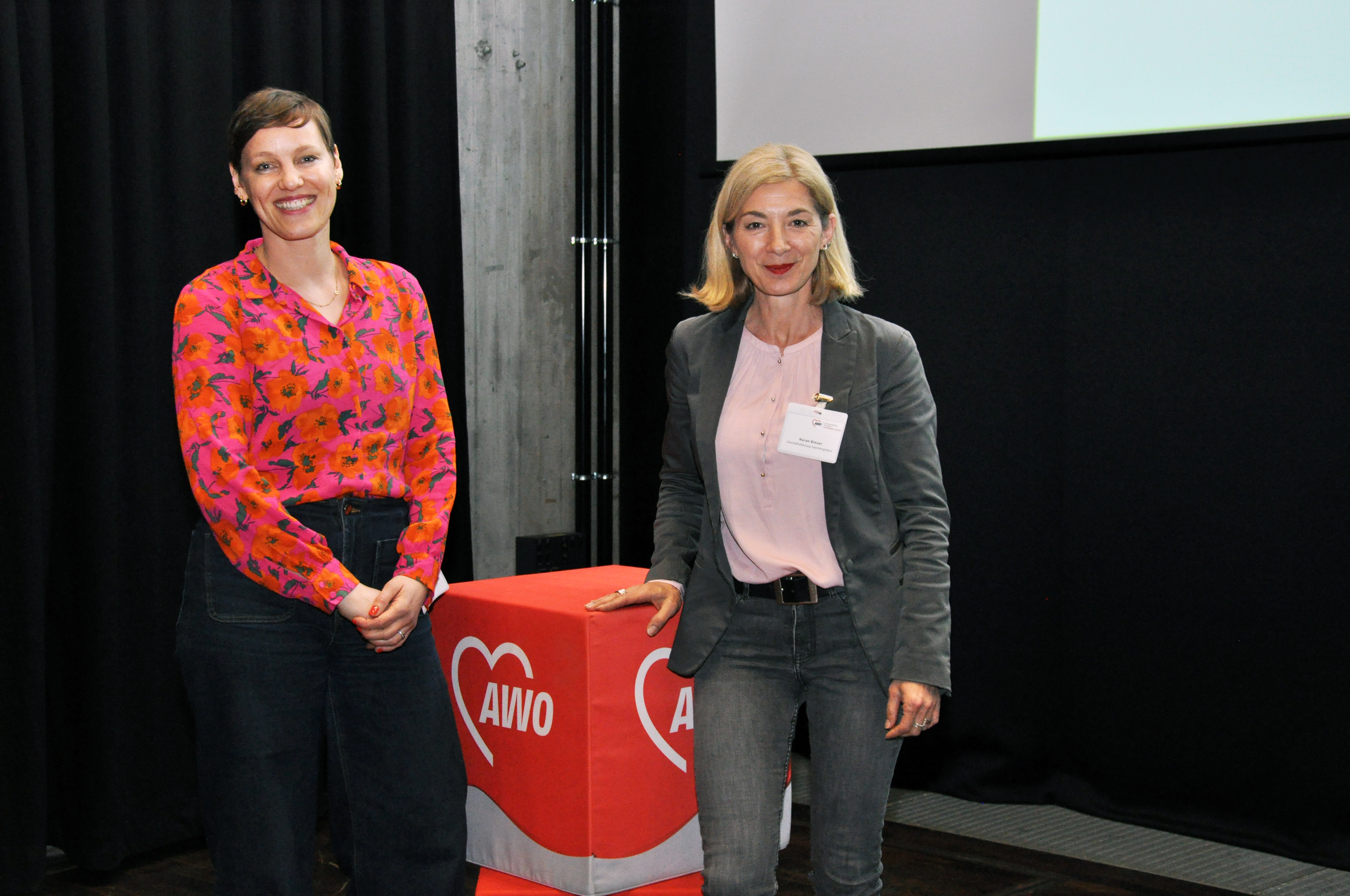 Prof*in Dr. Karla Verlinden und Faglo-Geschäftsführerin Nuran Breuer stehen neben einem Sitzwürfel mit AWO-Logo.