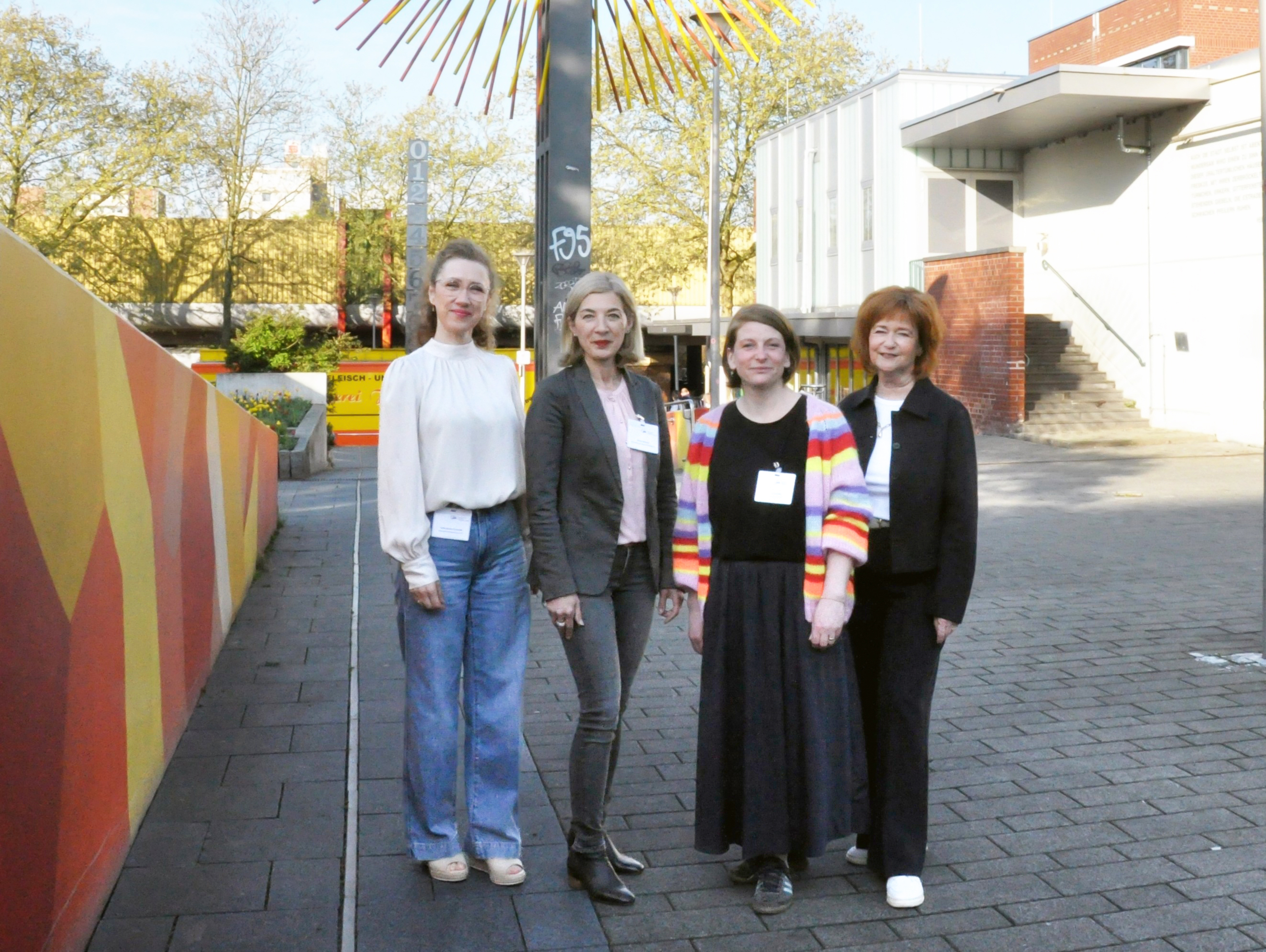 Gruppenfoto mit vier Personen neben einer bunt bemalten Wand, vor dem sogenannten "Sonnenrad", einer großen Skulptur, an der sich oben eine Art Sonne dreht.