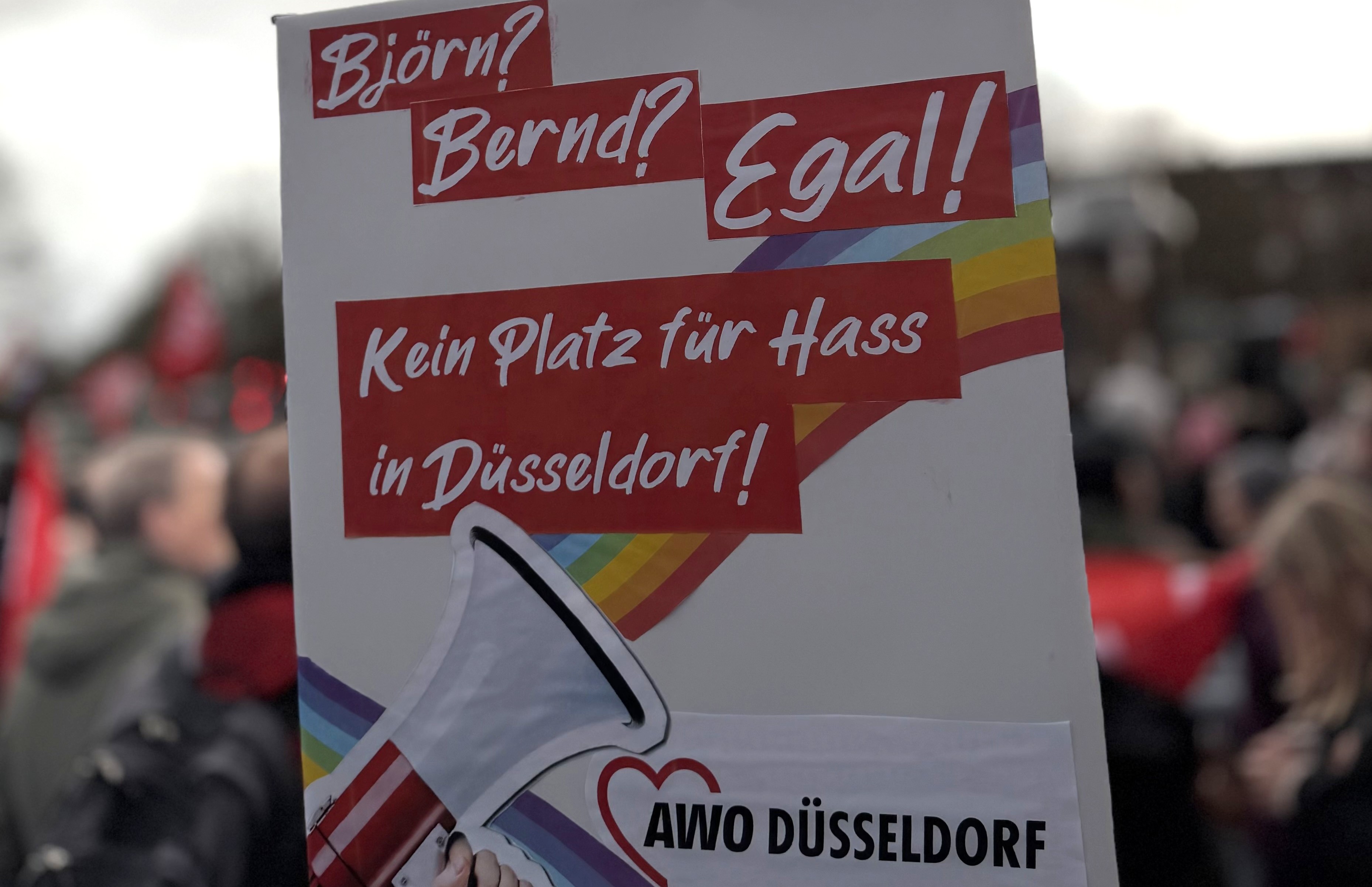 Foto von einem hochgehaltenen AWO-Plakat mit Megaphon, Regenbogen und dem Spruch: Björn? Bernd? Egal! Kein Platz für Hass in Düsseldorf. Im Hintergrund ist unscharf eine Menschenmenge zu sehen.